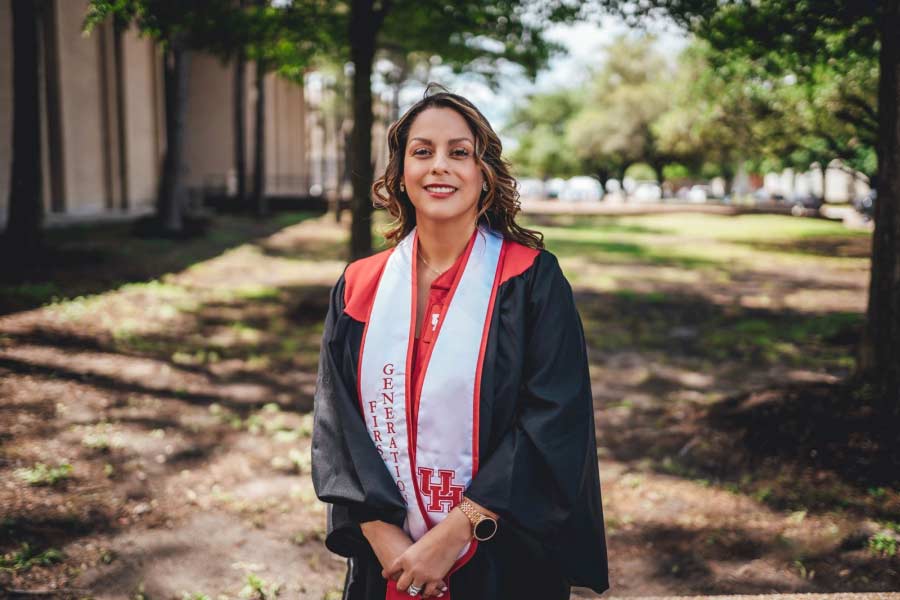 Portrait of a Hispanic woman smiling wearing graduation regalia with a white sash..