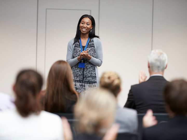 A smiling black woman in a gray dress and cardigan speaking in front of a room of seated individuals facing the speaker