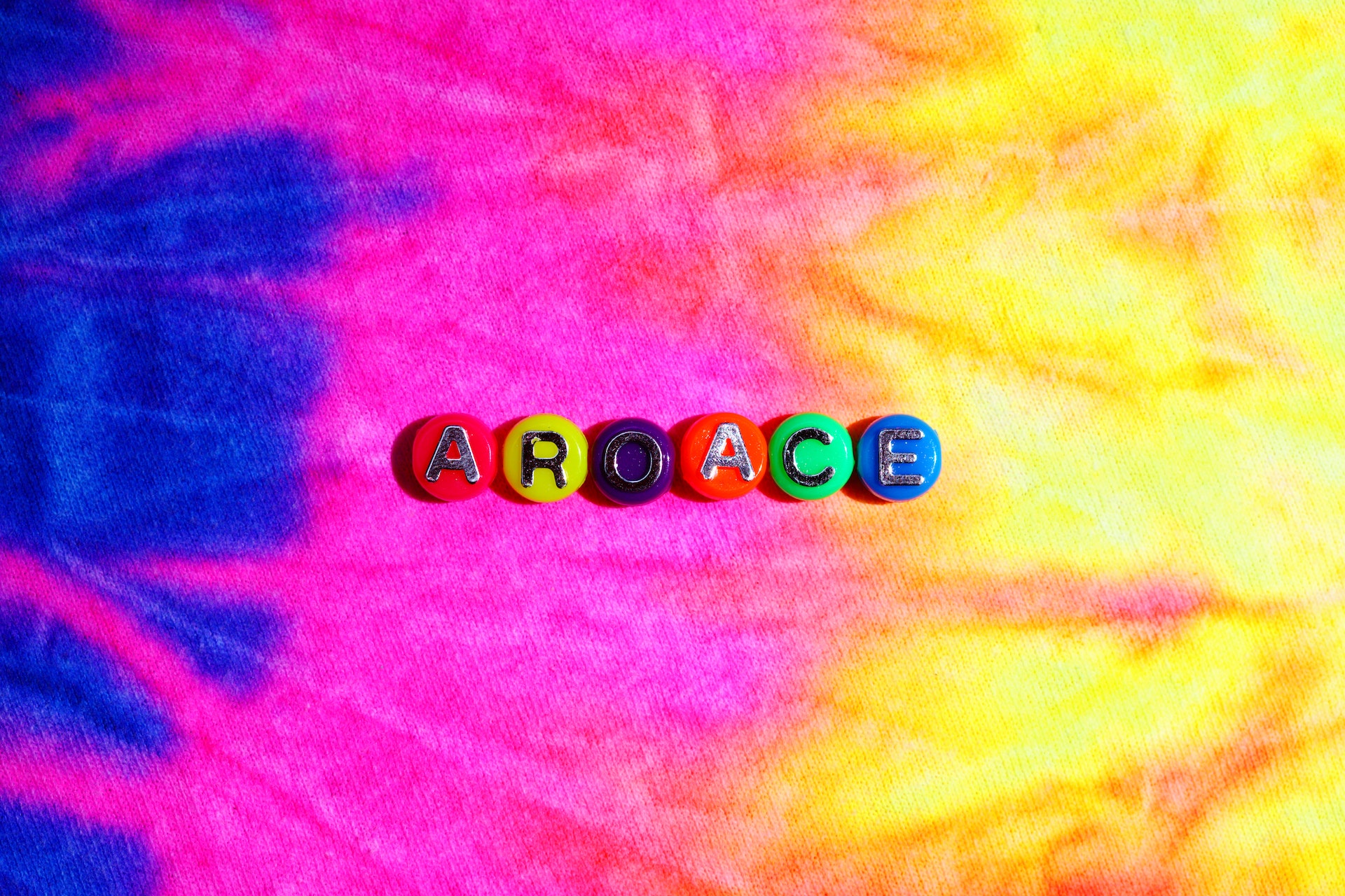 A
rainbow/tie-dye
background
with
beads
spelling
out
“ARO/ACE”.