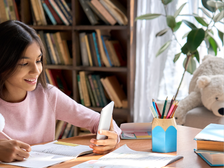 Girl in a pink sweater looking at her phone while sitting at her desk. Book shelf in the background