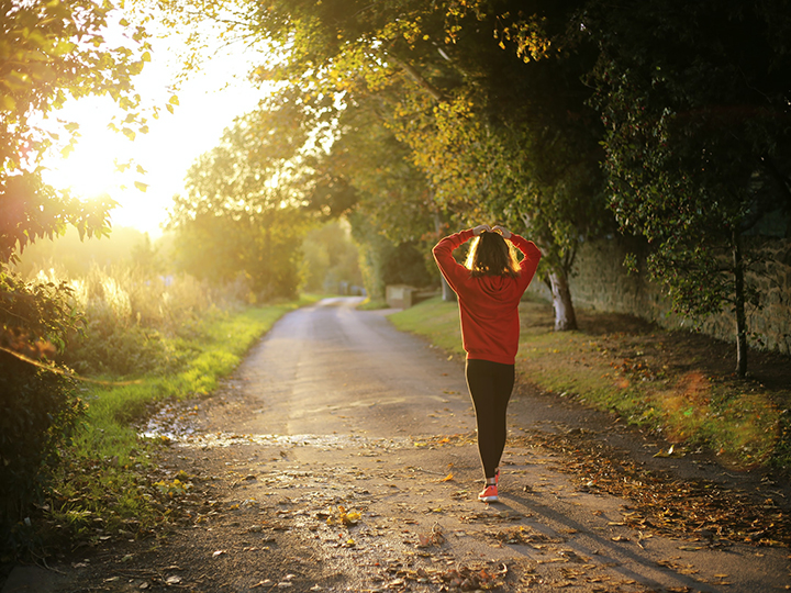A women in a red shirt walking down a tree-lined path in the sunlight.