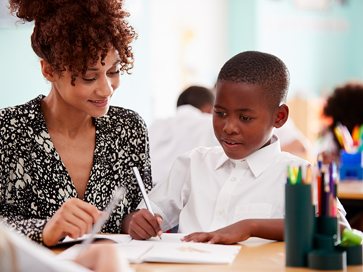 Woman Elementary School Teacher Giving Male Pupil Wearing Uniform One To One Support In Classroom