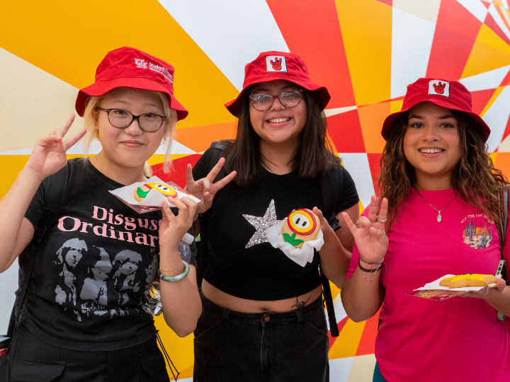 three female students holding up a mario themed sugar cookie while smiling to the camera wearing bucket hats