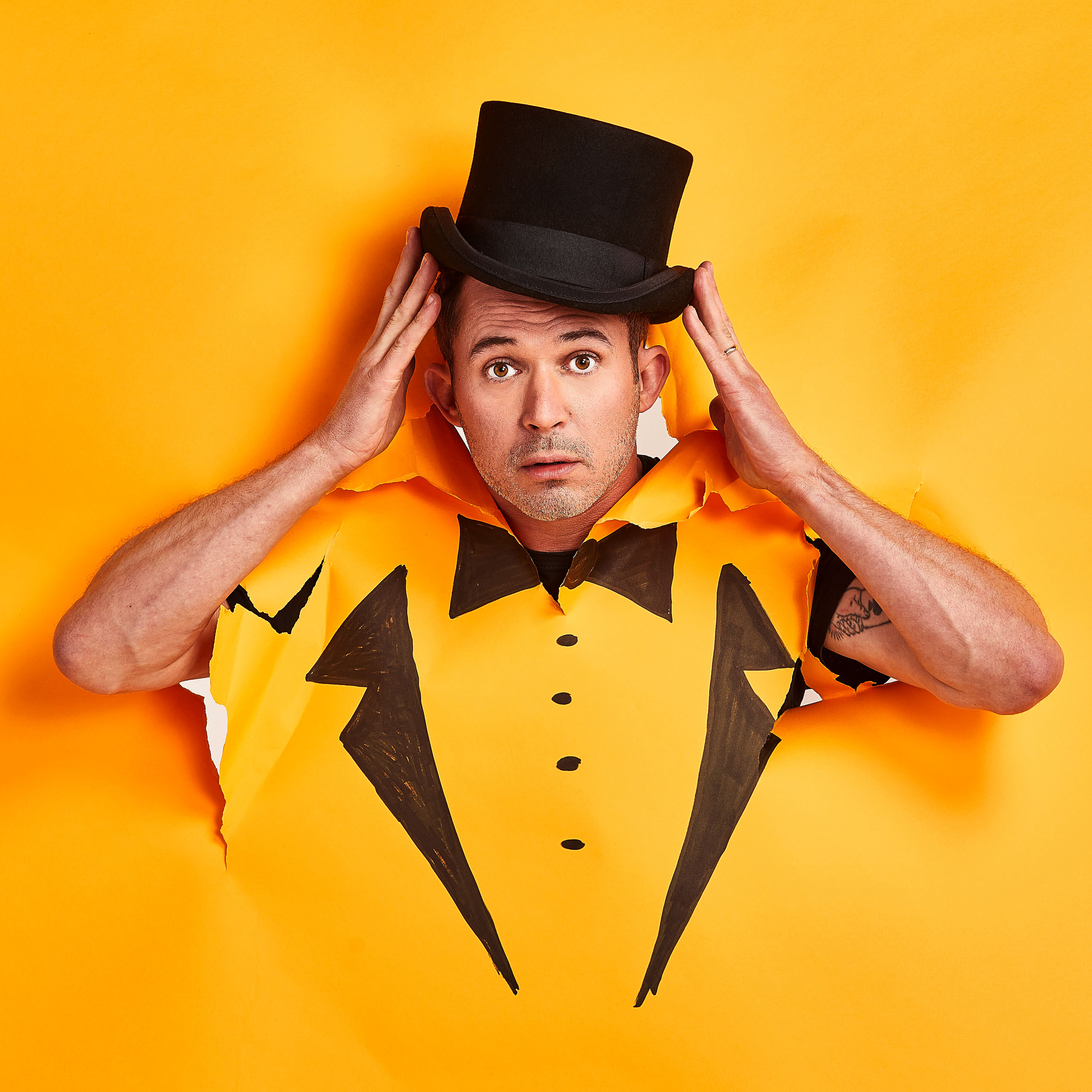 Justin Willman's head and arms pop out of a yellow paper background that has the illustration of a tux drawn on and colored in sharpie. His hands are up near his head holding a top hat on top of his head. 
