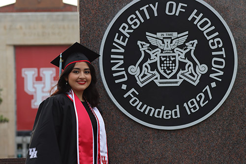 A woman with long brown hair in regalia standing in front of the UH administration building