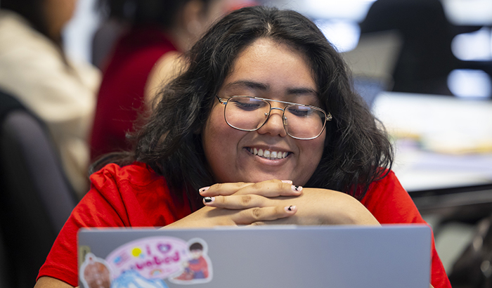 A student with brown hair and glasses looking at a laptop smiling