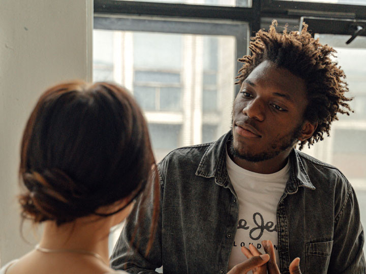 A Black man in a long sleeve gray shirt talking to a woman in a hallway