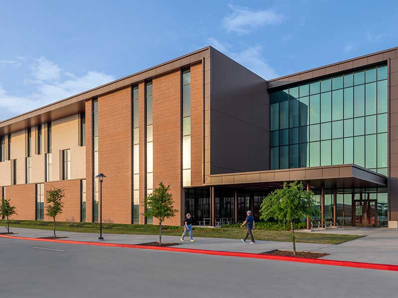 Photograph of a large three story brick and glass building with students walking on the sidewalk.