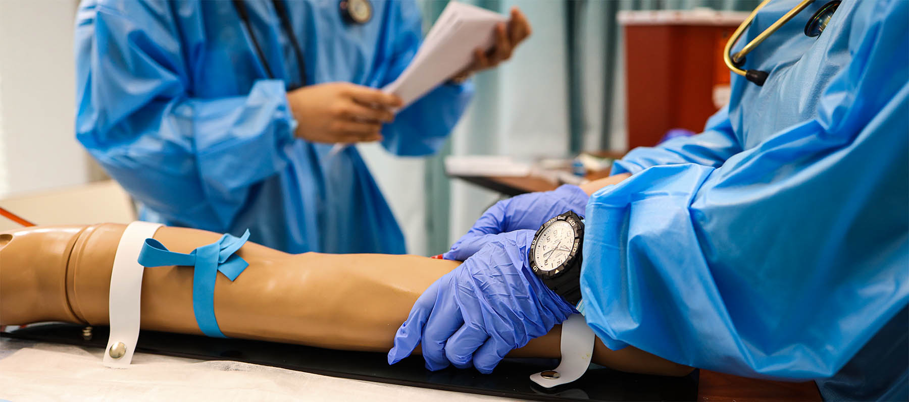 A closeup of a nursing student's hands with gloves holding a mannequin's arm that is wrapped in a tourniquet.