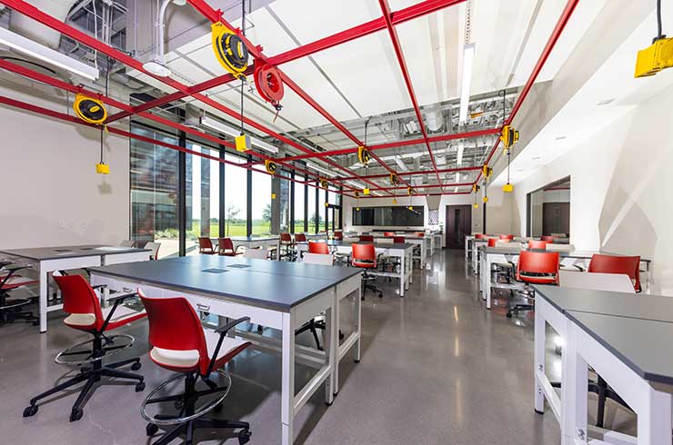 Photograph of an engineering lab with groups of desks with swivel chairs. A red metal frame with hanging outlets are over the desks.