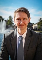 Portrait of dark blonde man in a suit in front of a lake with a fountain