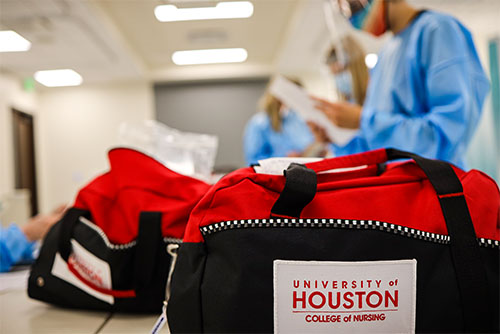 Close up of a black and red bag with the College of Nursing logo. Nursing students in scrubs can be seen in the background. 