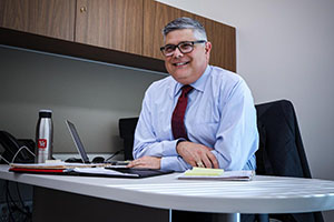 A man in a light blue dress shirt smiling while sitting at a desk in an office