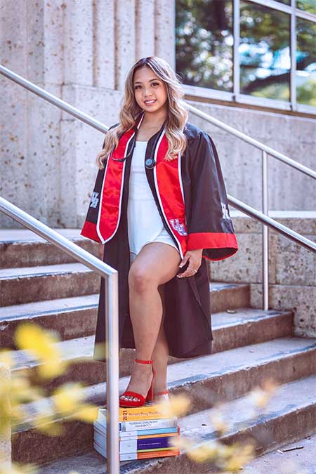 A hispanic woman dressed in black and red graduation regalia stands on outdoor concrete stairs.