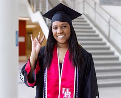 A black female woman dressed in black and red graduation regalia holds her right hand in the UH Cougar Paw gesture.