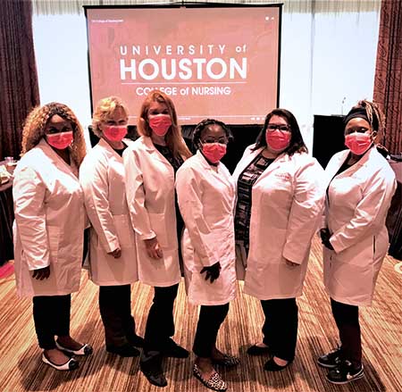 A group of women nurses in lab coats stand in front of a screen that says "University of Houston College of Nursing".