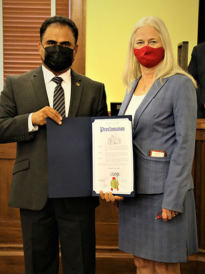 A man in a suit holds a black folder with a proclamation inside while standing next to a woman wearing a suit. Both are wearing face masks.