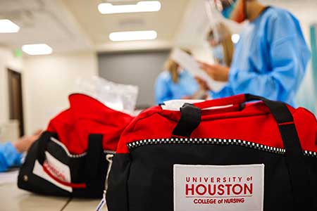 A black and red bag with the logo "University of Houston College of Nursing" is in the foreground. Students in blue protect gowns stand in the background.