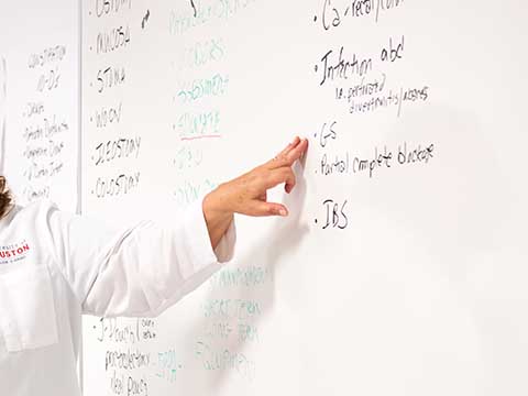 A faculty member in a white lab coat points to writing on a white board.