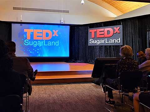 An image of a stage with a background that reads "TEDX Sugar Land."