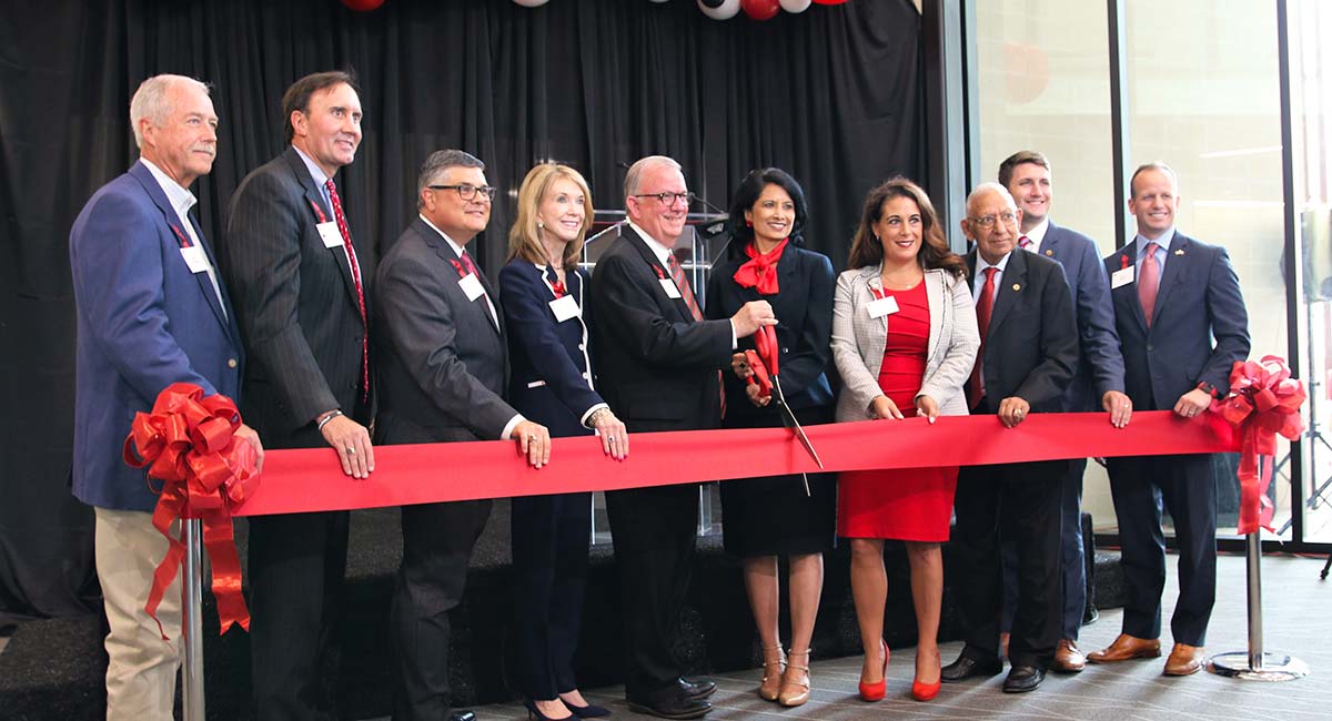 A group of men and women in business attire stand behind a ribbon hung from two stancions. The man and woman in the middle are holding an extra-large pair of scissors.