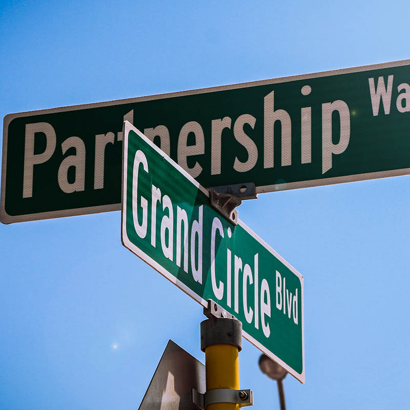 Street signs on a traffic sign pole. The top signs says "Partnership Way" and the lower one says "Grand Circle Blvd".