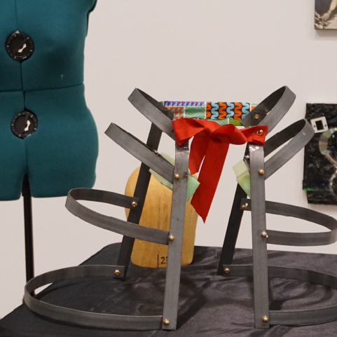 A dark-metal underskirt with two halves connected by a red bow resting on a black-cloth surface in front of a teal mannequin bust and smooth wooden mannequin head.