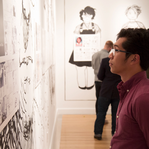 A person with short dark hair, wearing glasses and a maroon collared shirt stands in profile looking at artwork on the wall, other people stand around in the background.