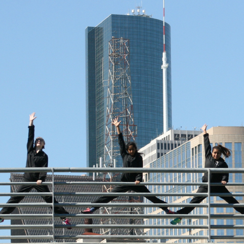 Performers in black coveralls simultaneously mid-split jump with one arm raised and the other holding railing atop a bridge in front of a downtown skyline.