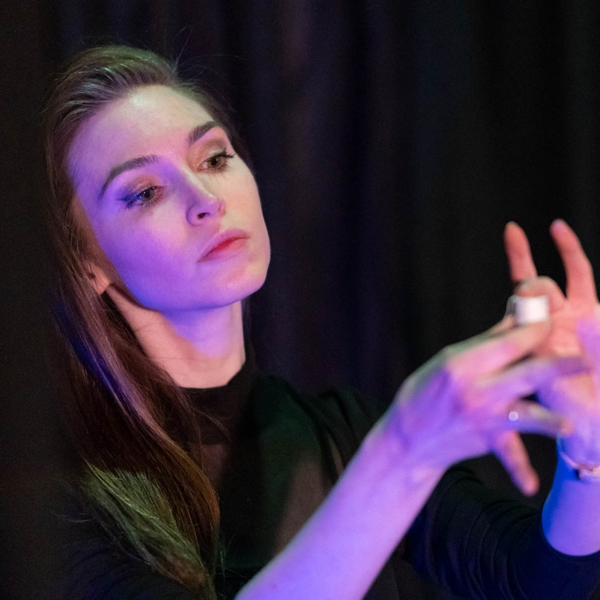 Illusionist and Artist Jeanette Andrews is light with a cool magenta light and performs a magic trick, holding a spool of white thread dramtically between her fingers 