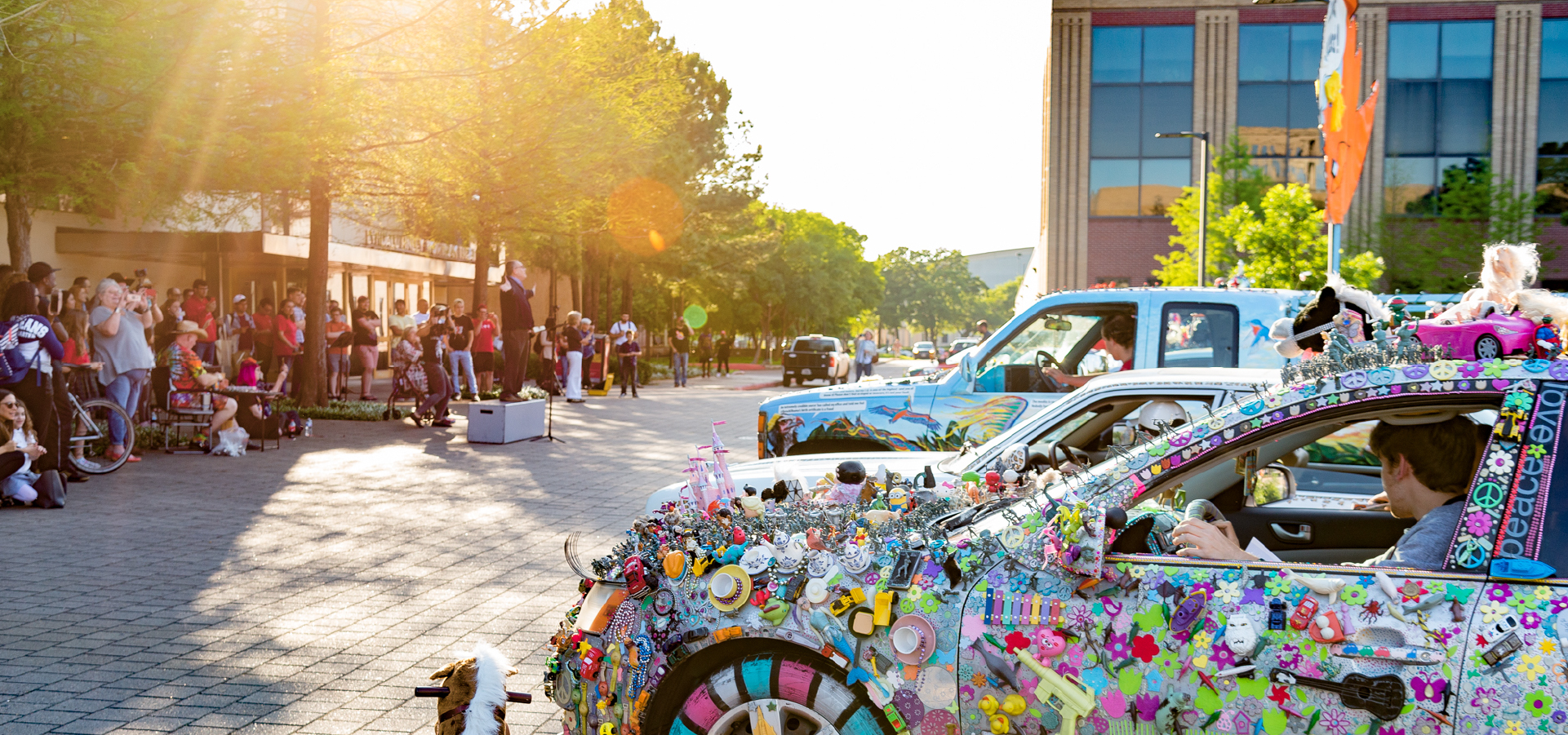 A crowd in front of the Mitchell Ceneter building stand behind a presenter facing several art cars displayed on the right which are decorated with paintings and toys and other paraphernalia.