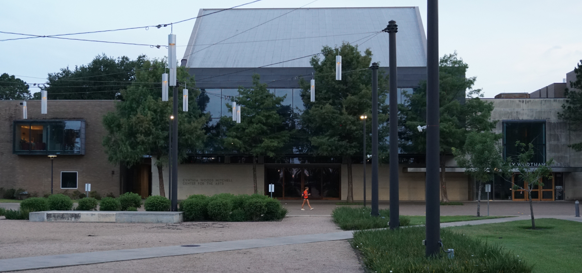 In the foreground, a grass and gravel courtyard with strung lighting design above; in the background a person wearing red walking by the Mitchell Center building during late afternoon.