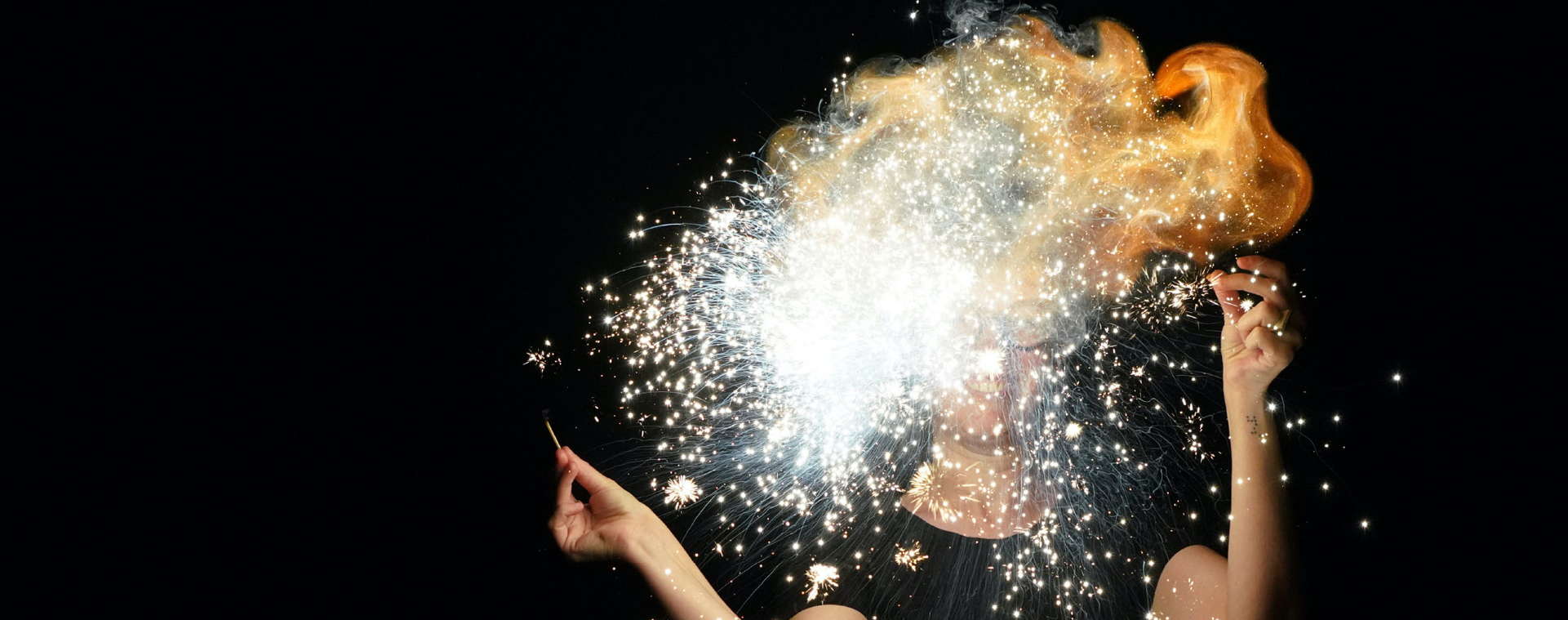 A close-up of a person’s face obscured by an explosion of sparks and fire against a black background.