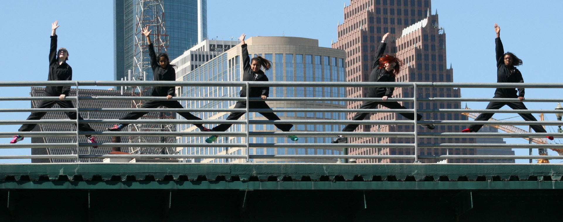 Performers in black coveralls simultaneously mid-split jump with one arm raised and the other holding railing atop a bridge in front of a downtown skyline.