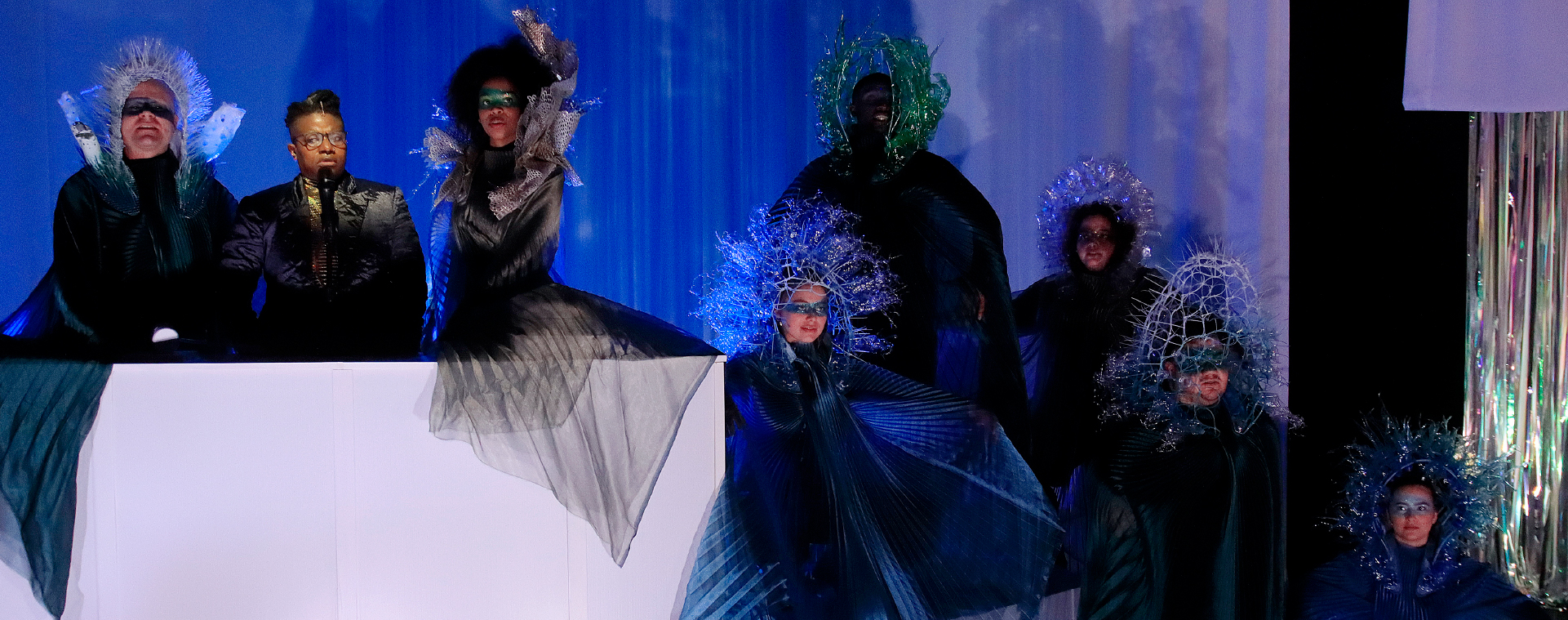 A performance with the main character speaking into a microphone behind a large podium surrounded by people in dark flowing costumes and elaborate headdresses in front of a blue backdrop.