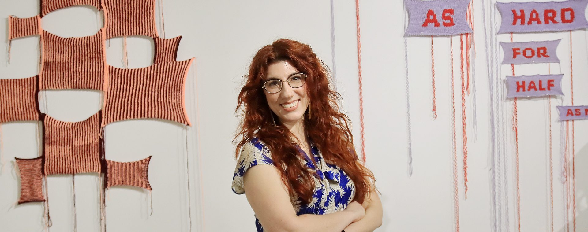 An artist with long red hair, wearing glasses and a blue patterned dress stands with arms crossed in front of a white wall with crocheted artwork that reads ‘Twice as hard for half as … ’ 