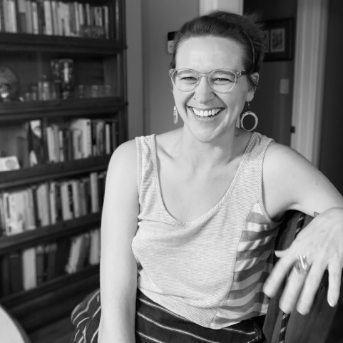 Black and white photo of a laughing, white-skinned woman with dark hair wearing glasses and hooped earrings sitting in a room with a full bookcase in the background.