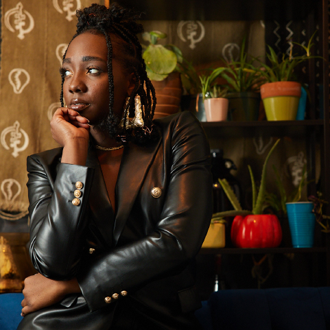 A black-skinned woman with black hair styled atop her head wearing a black coat, sits in front of a patterned wall and shelving full of plants with her head in her hand looking away.