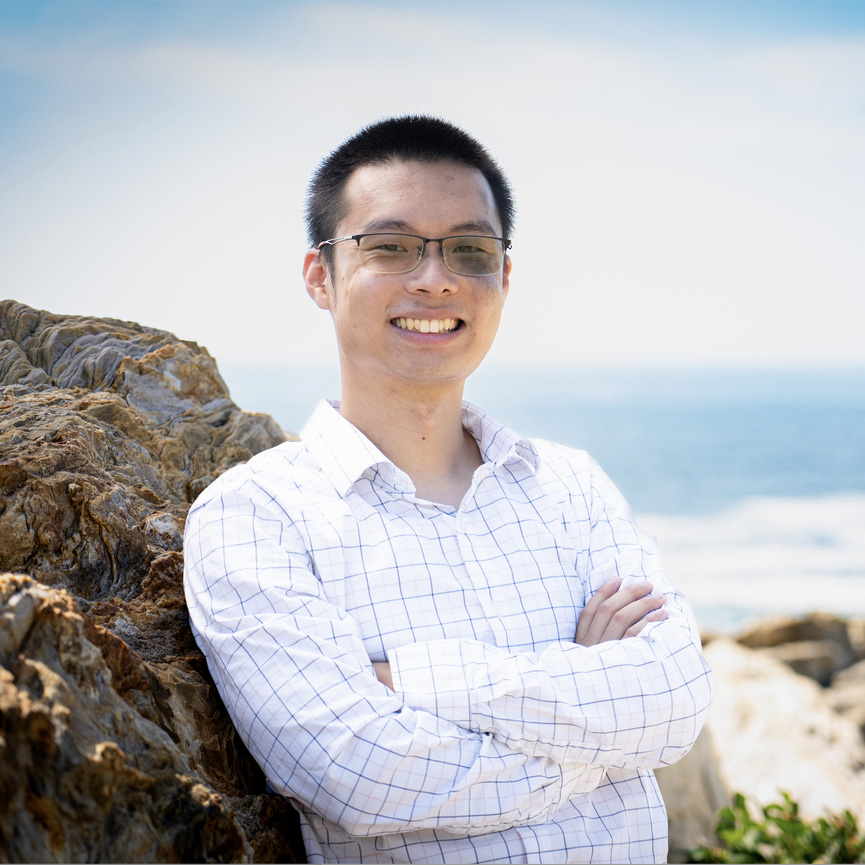 An Asian man poses with his arms crossed against his chest, wearing a plaid shirt, glasses, and smiling in front of rocks on a beach