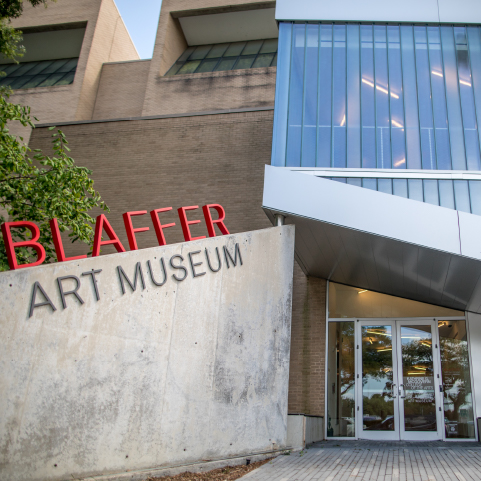 A concrete wall to the left with “Blaffer art museum” in red and grey letters leads to large glass entrance with a metal and glass second-floor above.