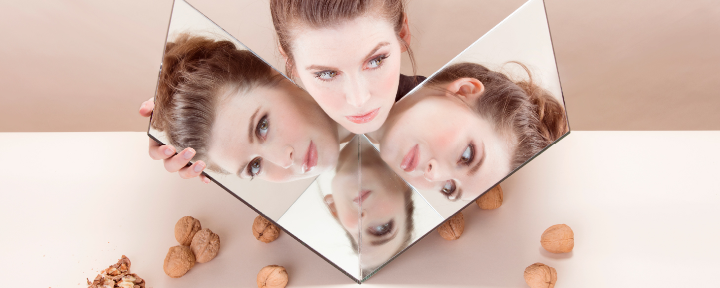 Jeanette Andrews’s face is reflected in multiple angles within a V-shaped mirror she holds under her chin resting on a white table scattered with walnuts against a peach-colored background.