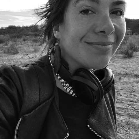 A black and white close-up of a smiling, light-skinned woman with dark hair wearing a dark jacket and headphones hanging around her neck. A desert-like landscape behind her.