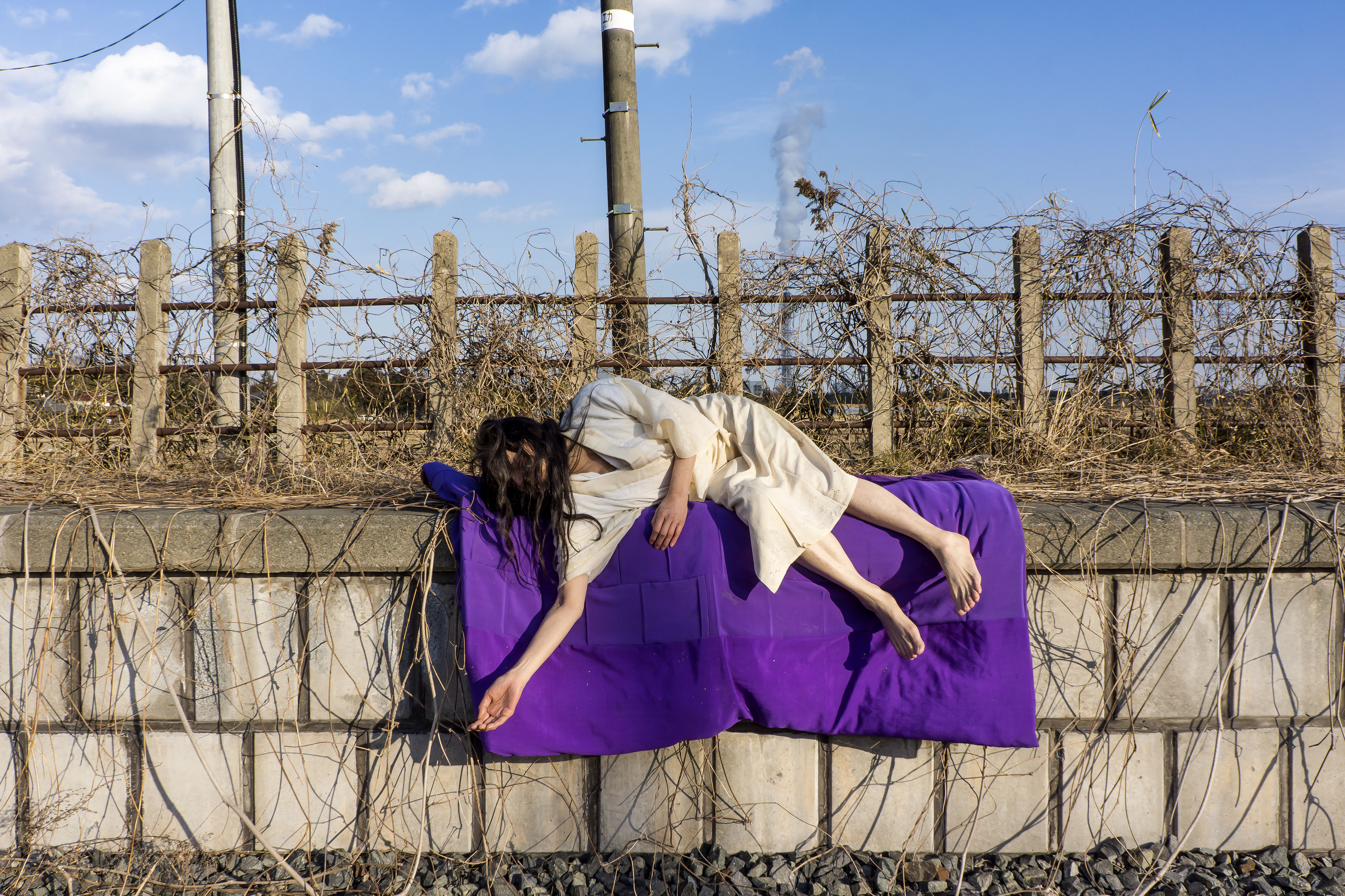 The artist lays draped in an industrial setting of concete with a purple textile underneath her body. She is wearing an off white dress and her body looks limp. 