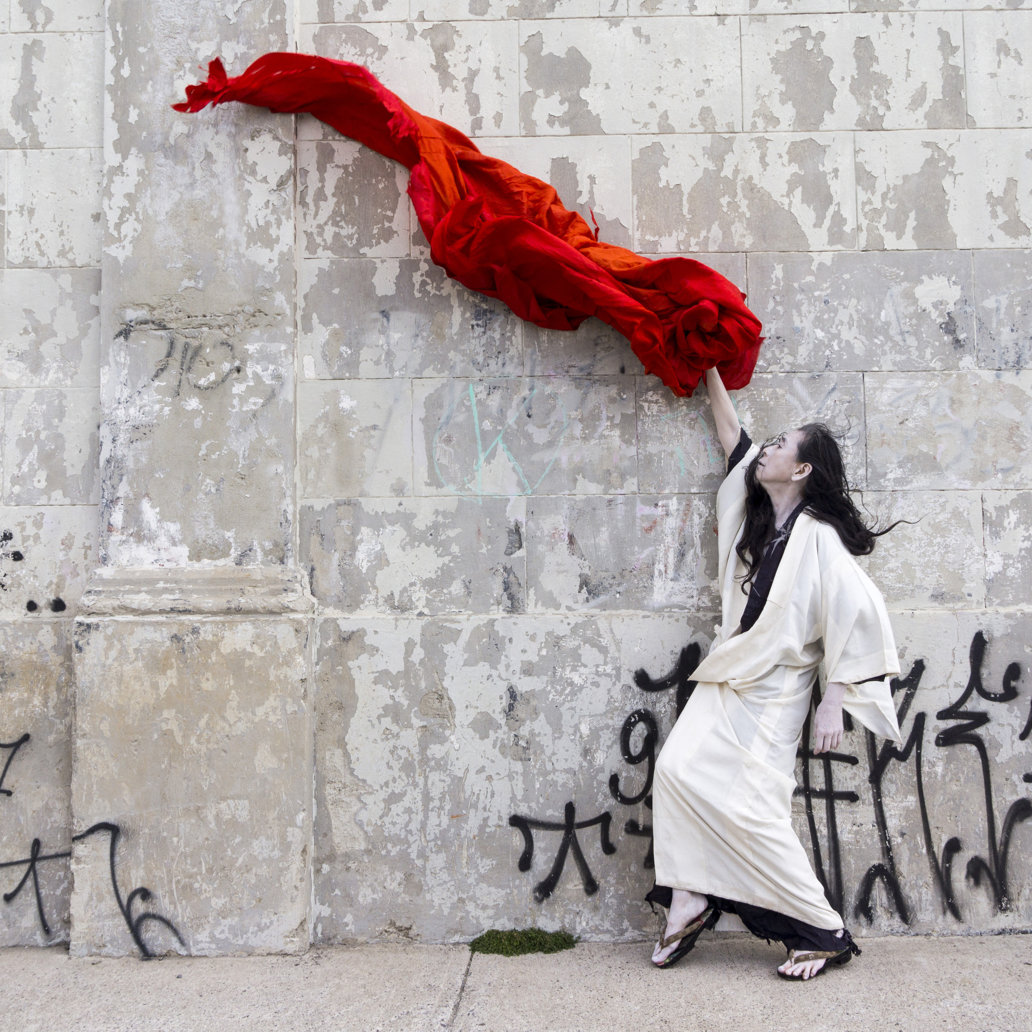 A japanese woman, slim and lithe, poses lyrically in front of an urban cement wall with a bright red textile