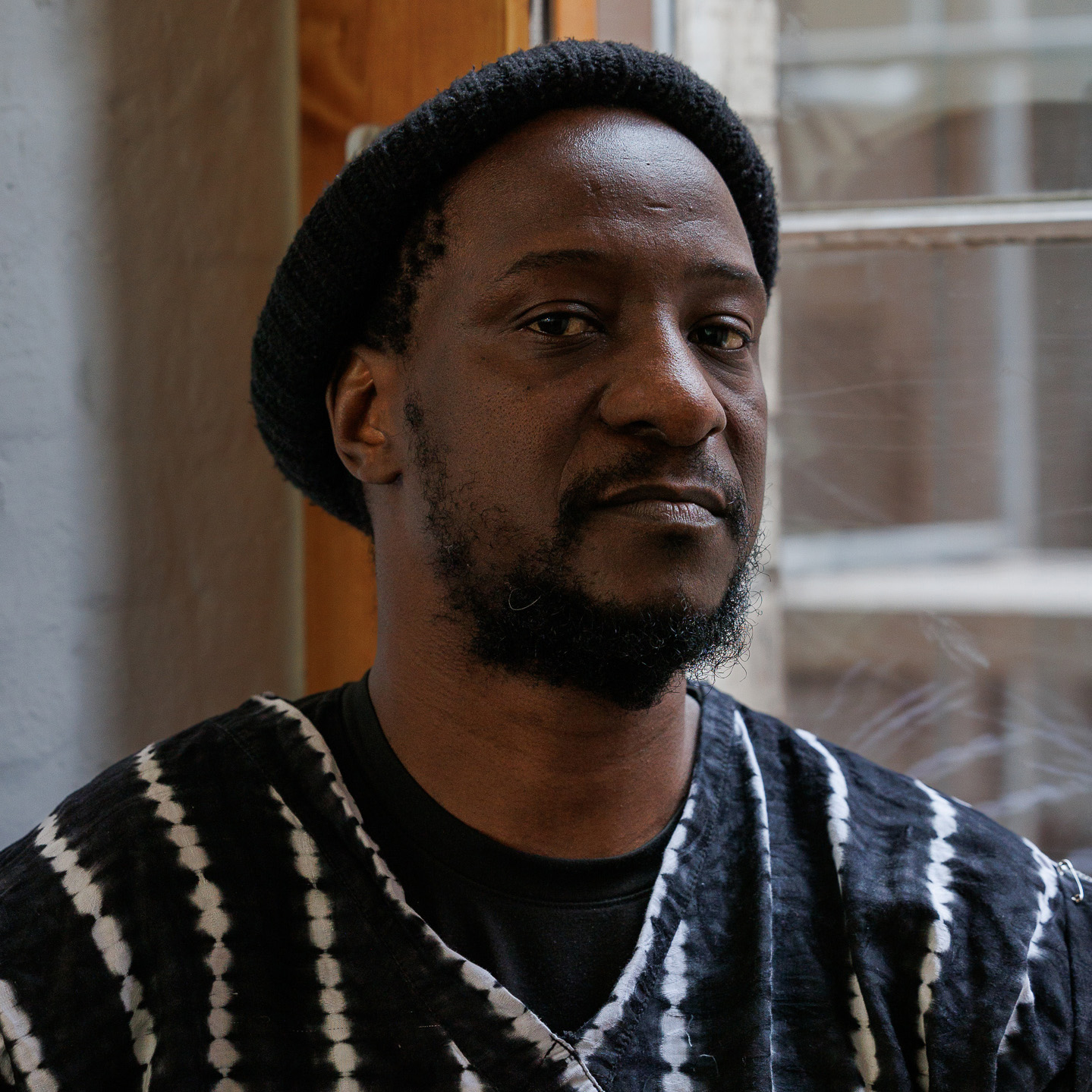A black man, slightly smiling, wearing a beanie in front of a window