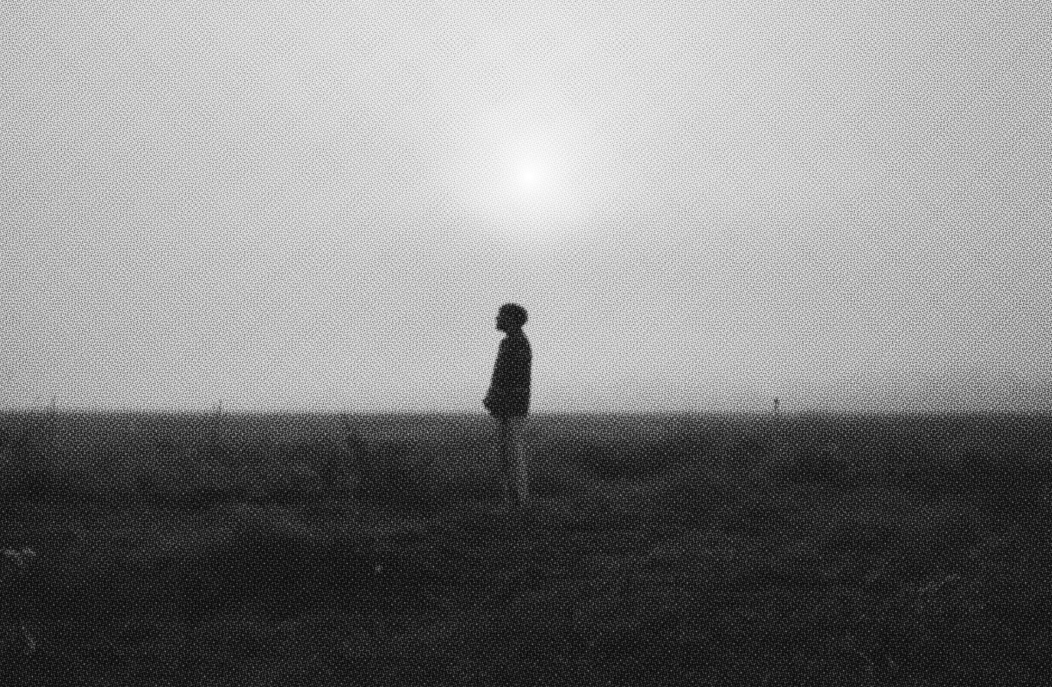 A person stands in alignment with the sun on a bleak, flat landscape. The photo is black and white and has a grainy treatment reminiscinet of vintage film. 