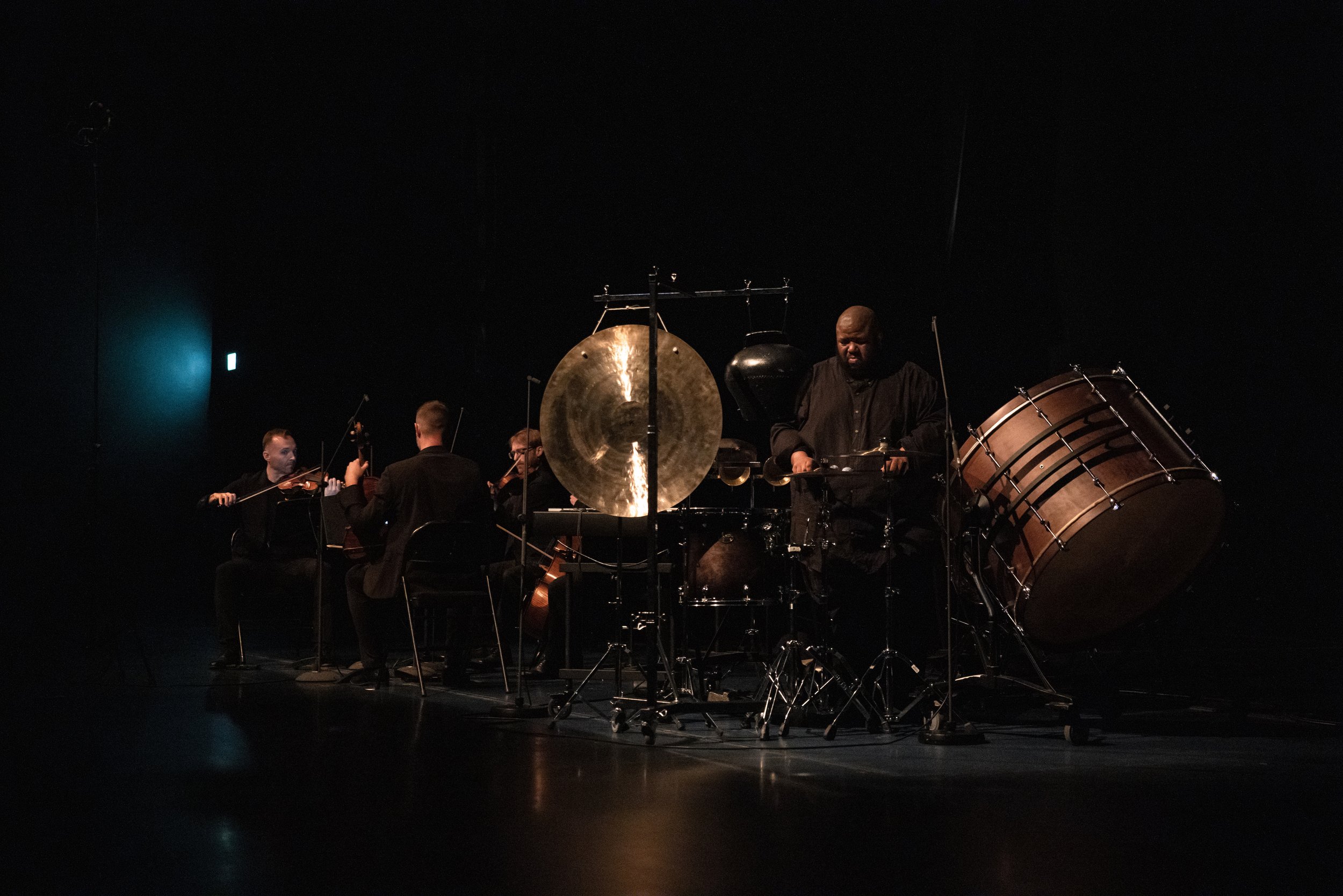 Tyshawn Sorey sits surrounded by percussion instruments such as cymbals performing music. Other musicians fill the stage around him. 