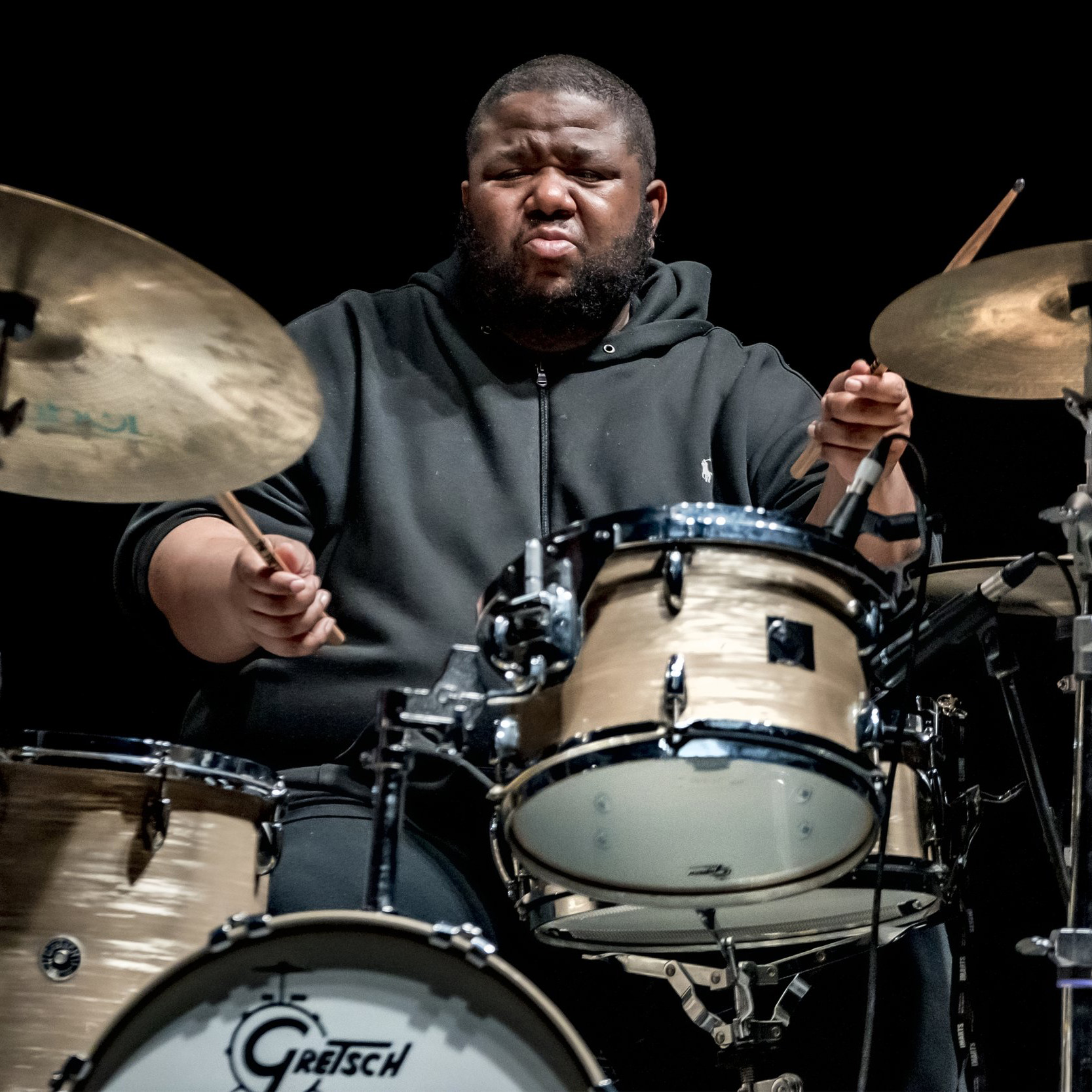 A black man sits at a drumset with a focused look on his face as he plays the drums