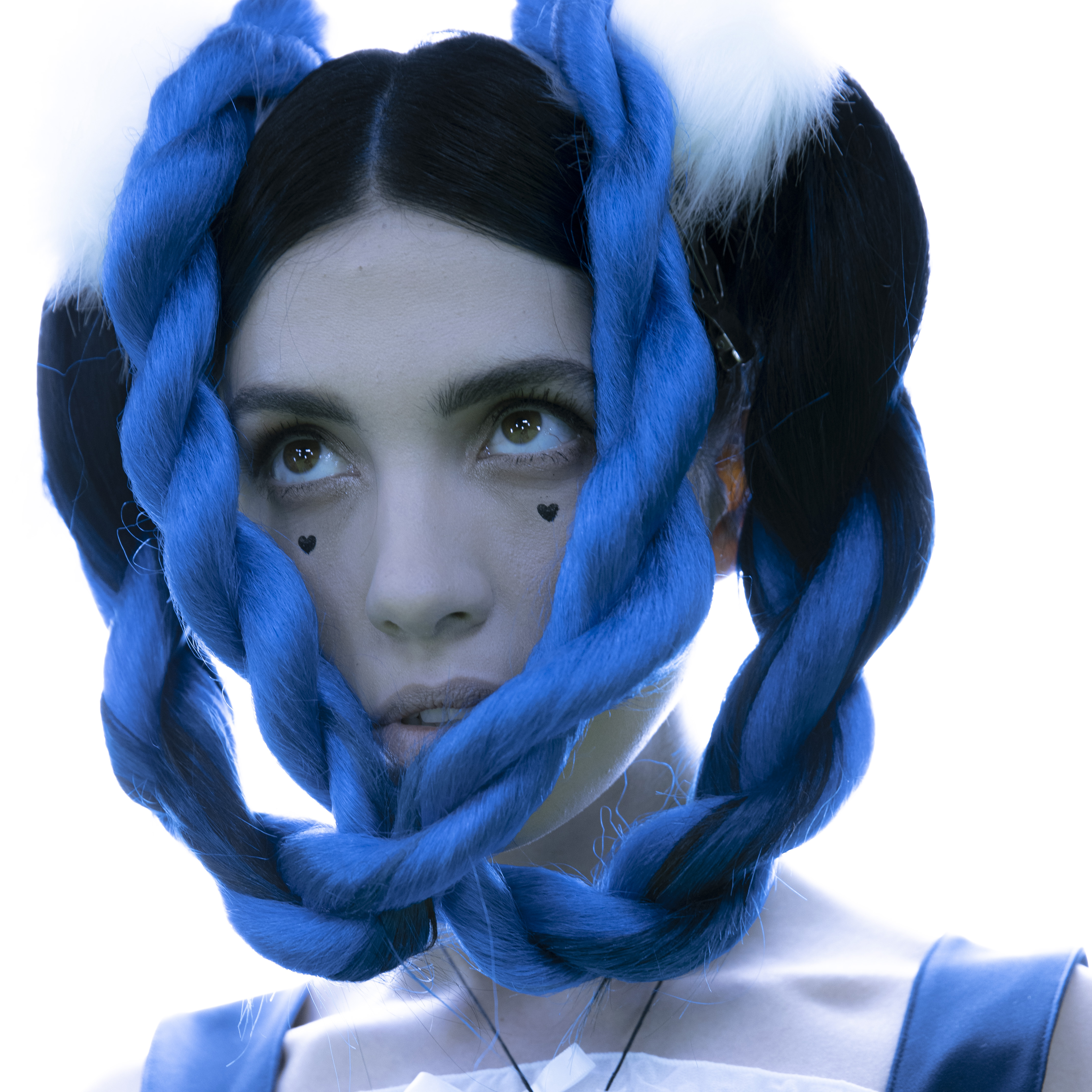 a Russian woman looks off away from the camera. she has dark brown and bright blue hair. the parts of her hair that are blue are twisted into braid like pigtails that cascade in front of her face. she has two black hearts under her eyes. she looks to be in her mid thirties. 