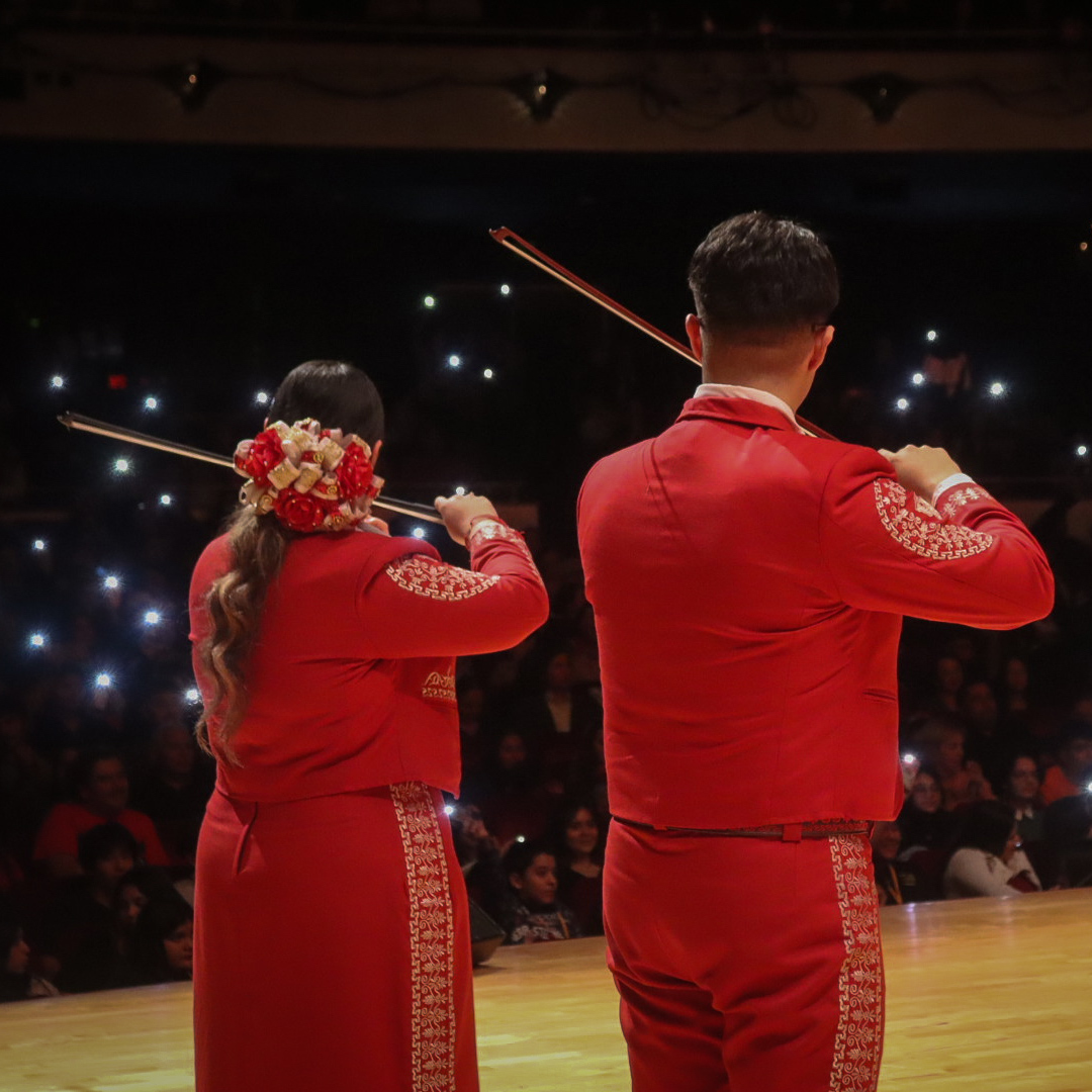 Mariachi violinists performing with audience members holding up phone lights in the background
